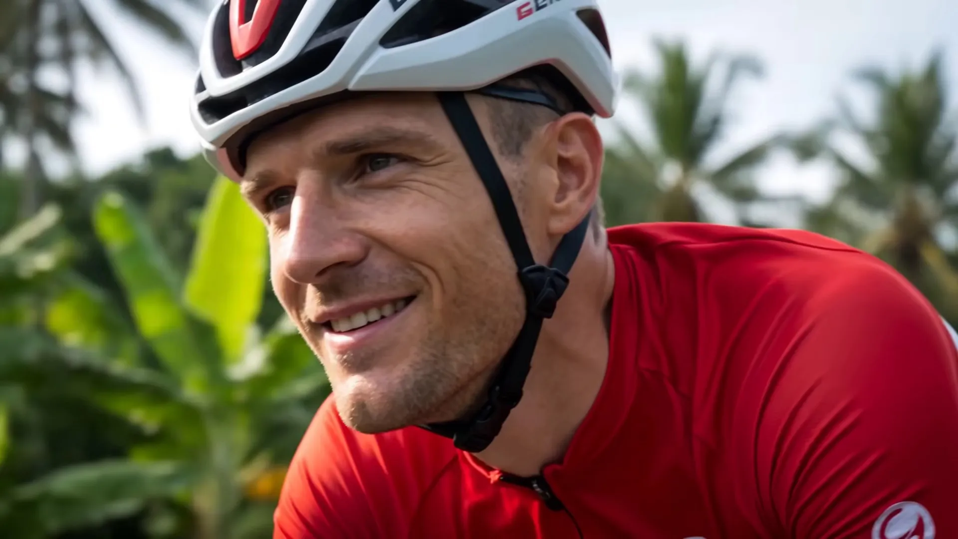A cyclist in a red jersey and white helmet riding through tropical highland country in bright daylight, banana palms blurred behind him.