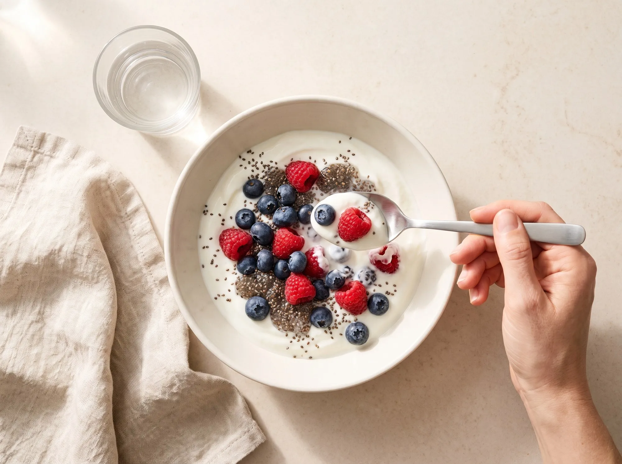 Seen from overhead on a pale stone counter in morning light: a shallow white bowl of thick yogurt topped with blueberries, raspberries and a scattering of dark chia seeds, a hand entering from the right lifting a spoonful, a linen napkin and a glass of water beside it.