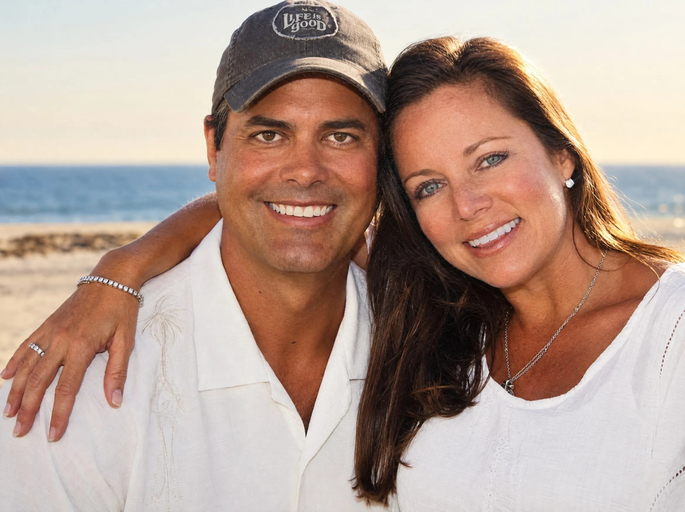 A smiling couple cheek to cheek on a beach in warm late-afternoon light: he in a grey cap and a white embroidered shirt, her arm around his shoulders; she with long brown hair in a white top, the ocean and sand soft behind them.