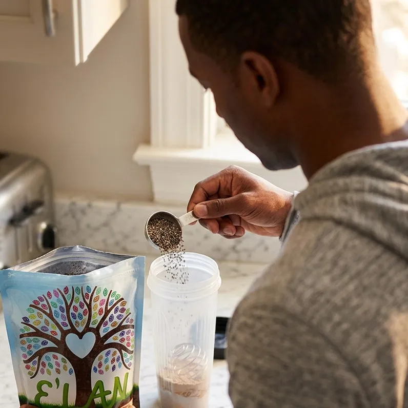 A man at a marble kitchen counter, seen from behind over his shoulder, tipping a measuring spoon of dry chia seed into a clear shaker bottle, with an open E'LAN pouch standing beside it.