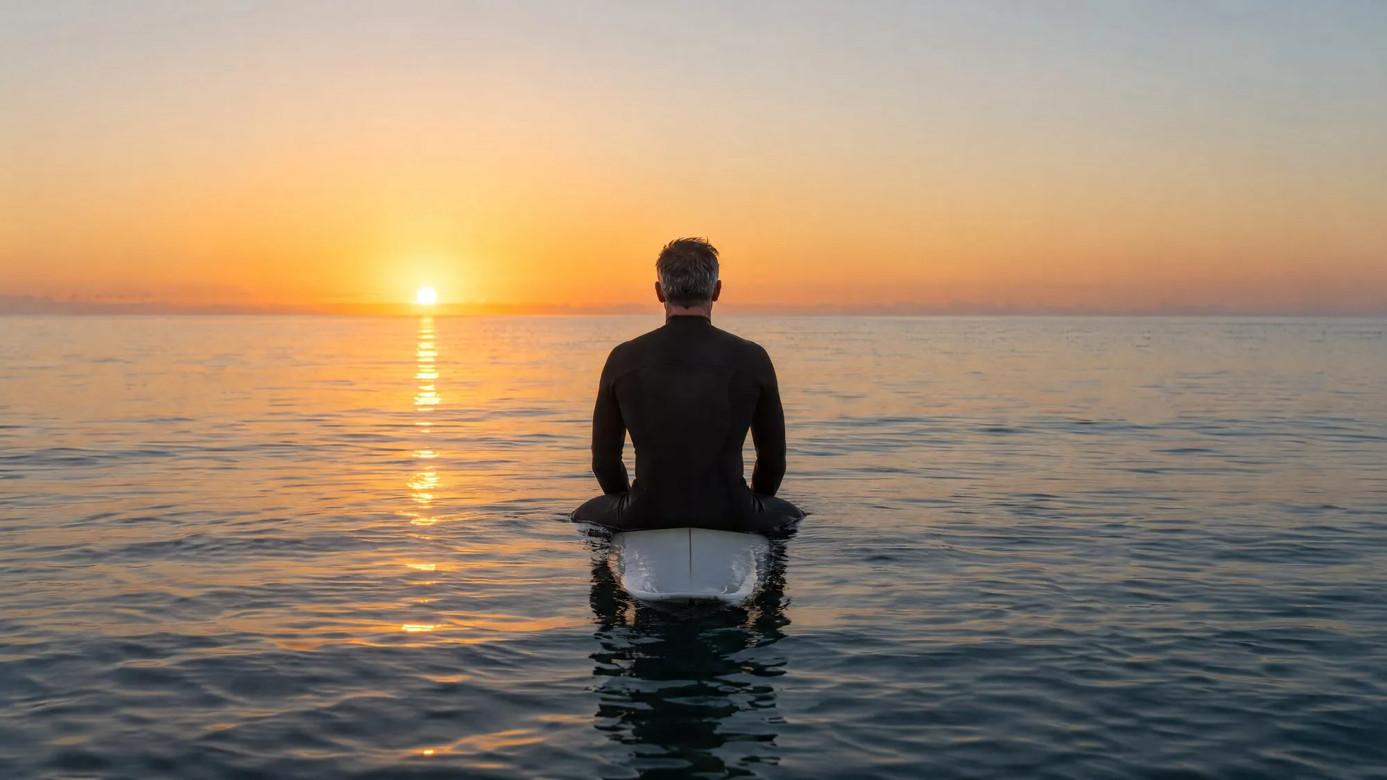 A grey-haired man in a black wetsuit sits on a surfboard on calm water, his back to the camera, watching the sun rise over the sea.